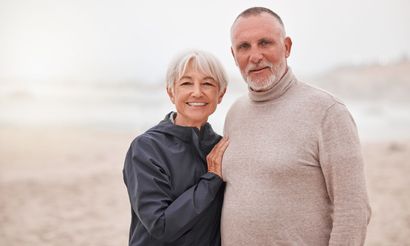 Smiling couple standing close together on a beach, wearing neutral-toned sweaters.