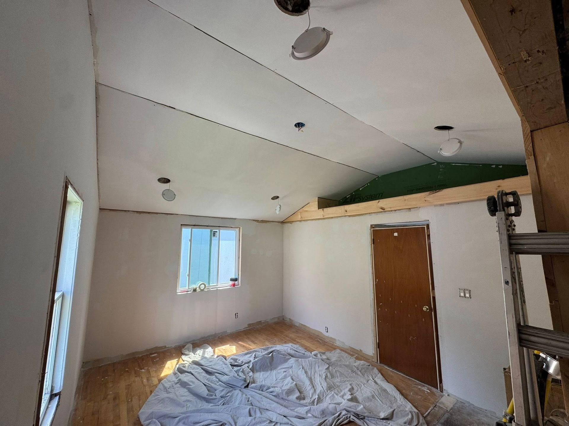 Partially finished room with white walls, sloped ceiling, wooden door, window, and a tarp-covered pile on the floor