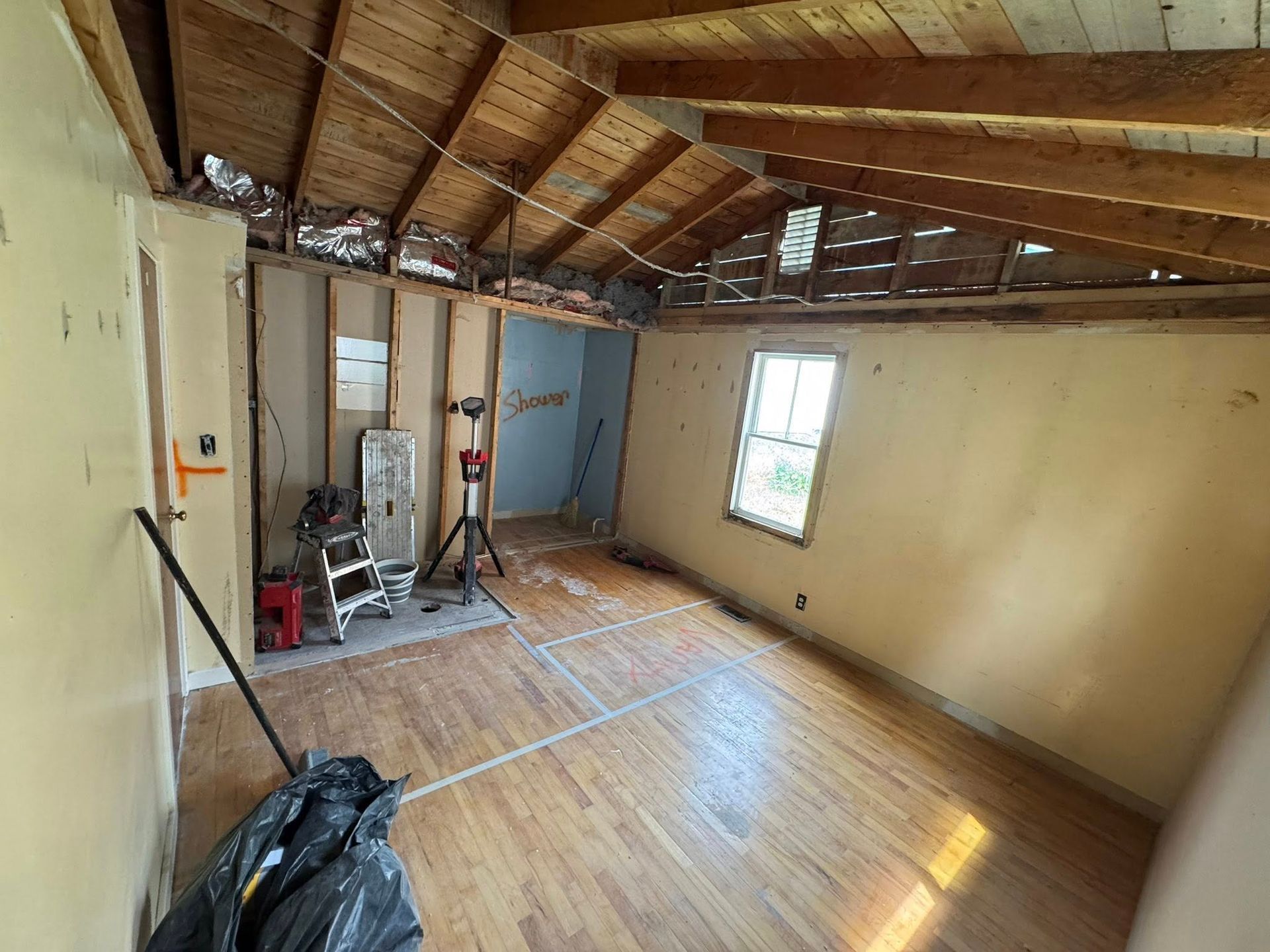 Empty room under renovation with exposed wooden ceiling, bare walls, and sunlight through a window.