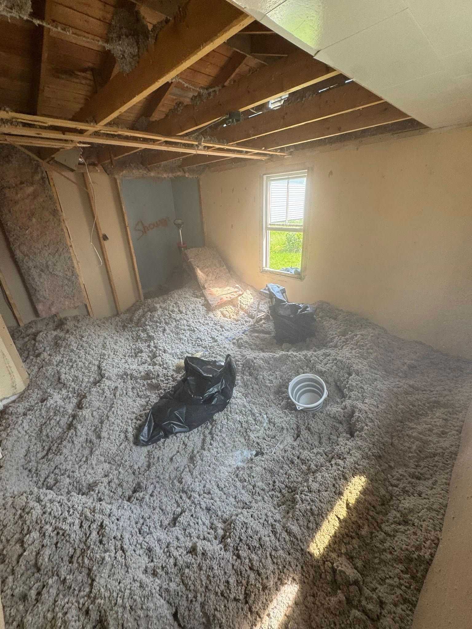 Attic room with exposed beams, white insulation, and a window letting in sunlight.