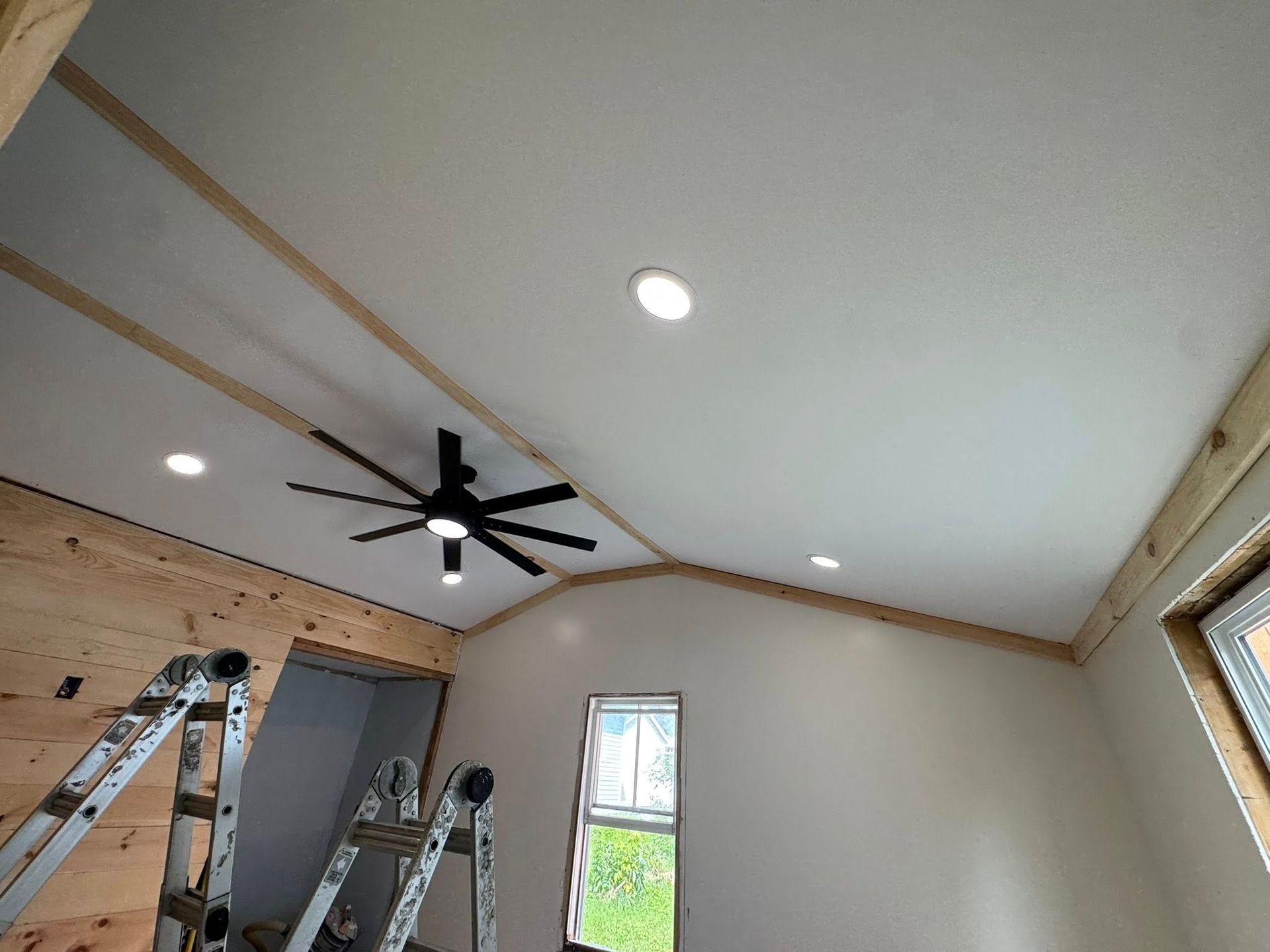 Room under renovation with recessed lights, black ceiling fan, ladders, and exposed trim on a vaulted ceiling