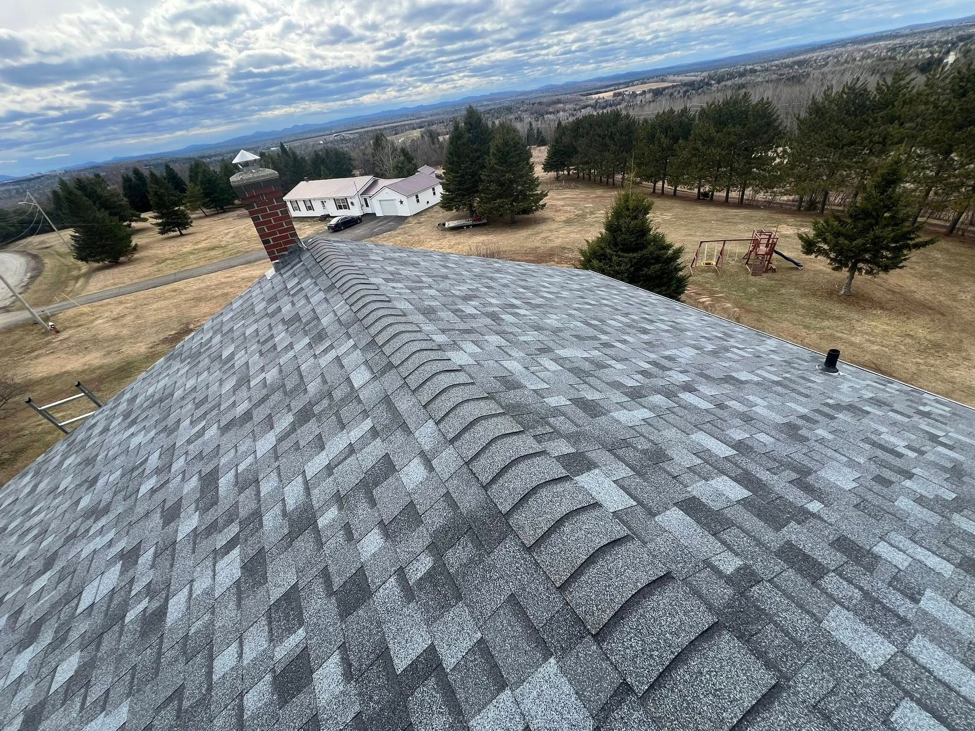 Gray shingled roof overlooking a rural landscape with trees and a cloudy sky