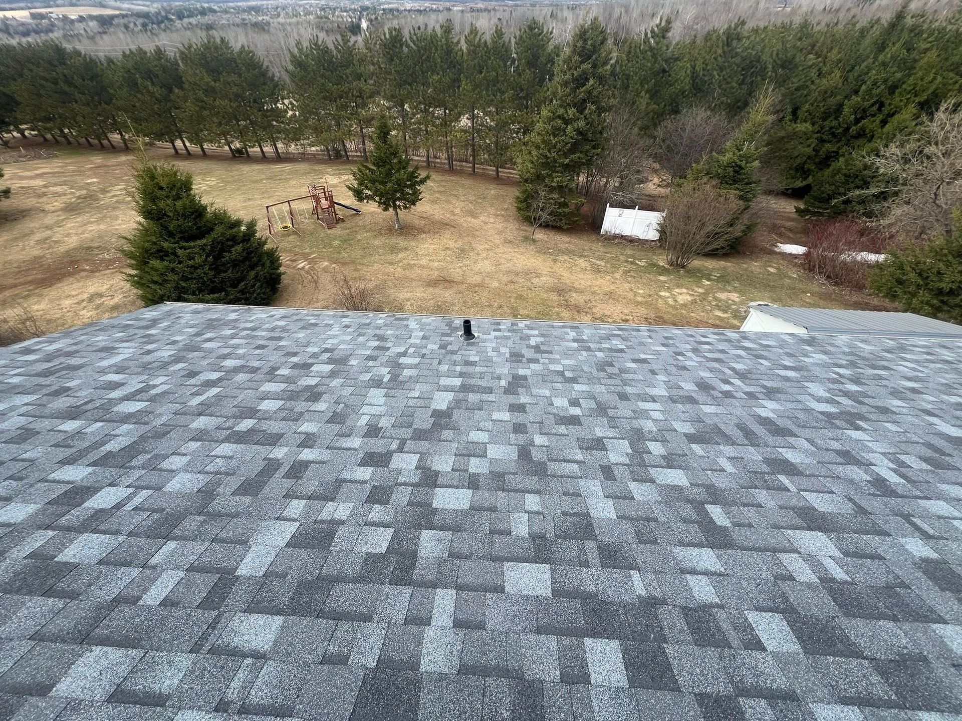 Gray shingled rooftop overlooking a grassy yard with trees under an overcast sky.
