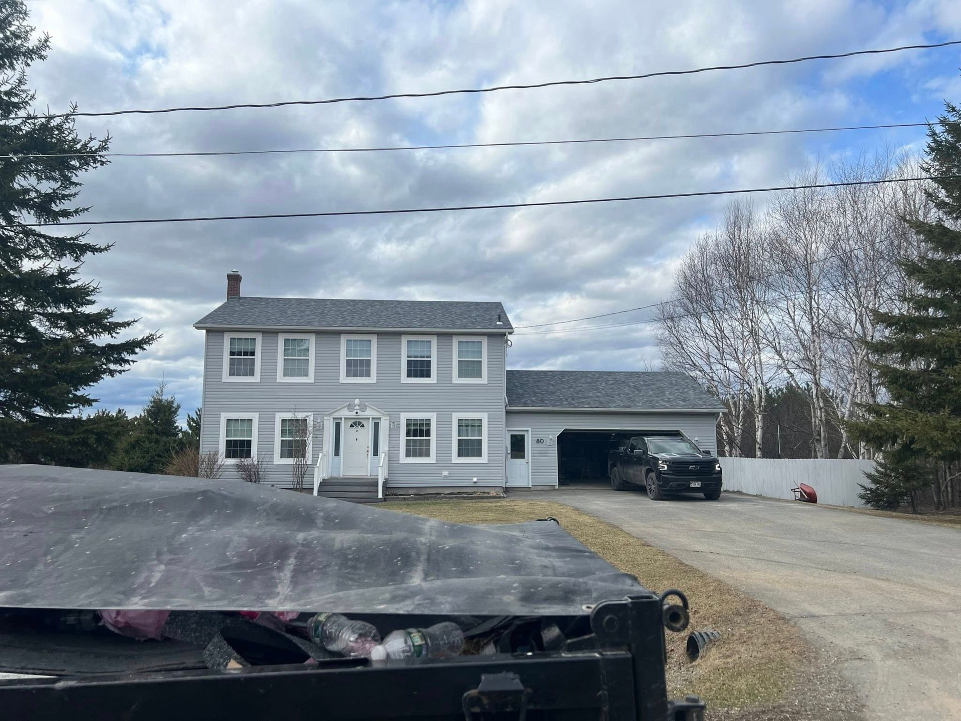 Gray two-story house with attached carport, gravel driveway, and cloudy sky behind a parked truck bed in foreground