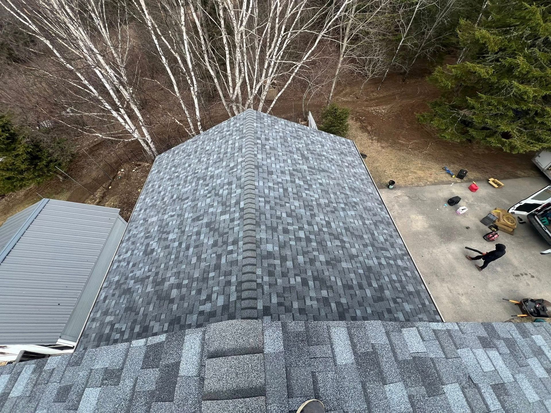Aerial view of a shingled roof beside a driveway and bare trees in a yard