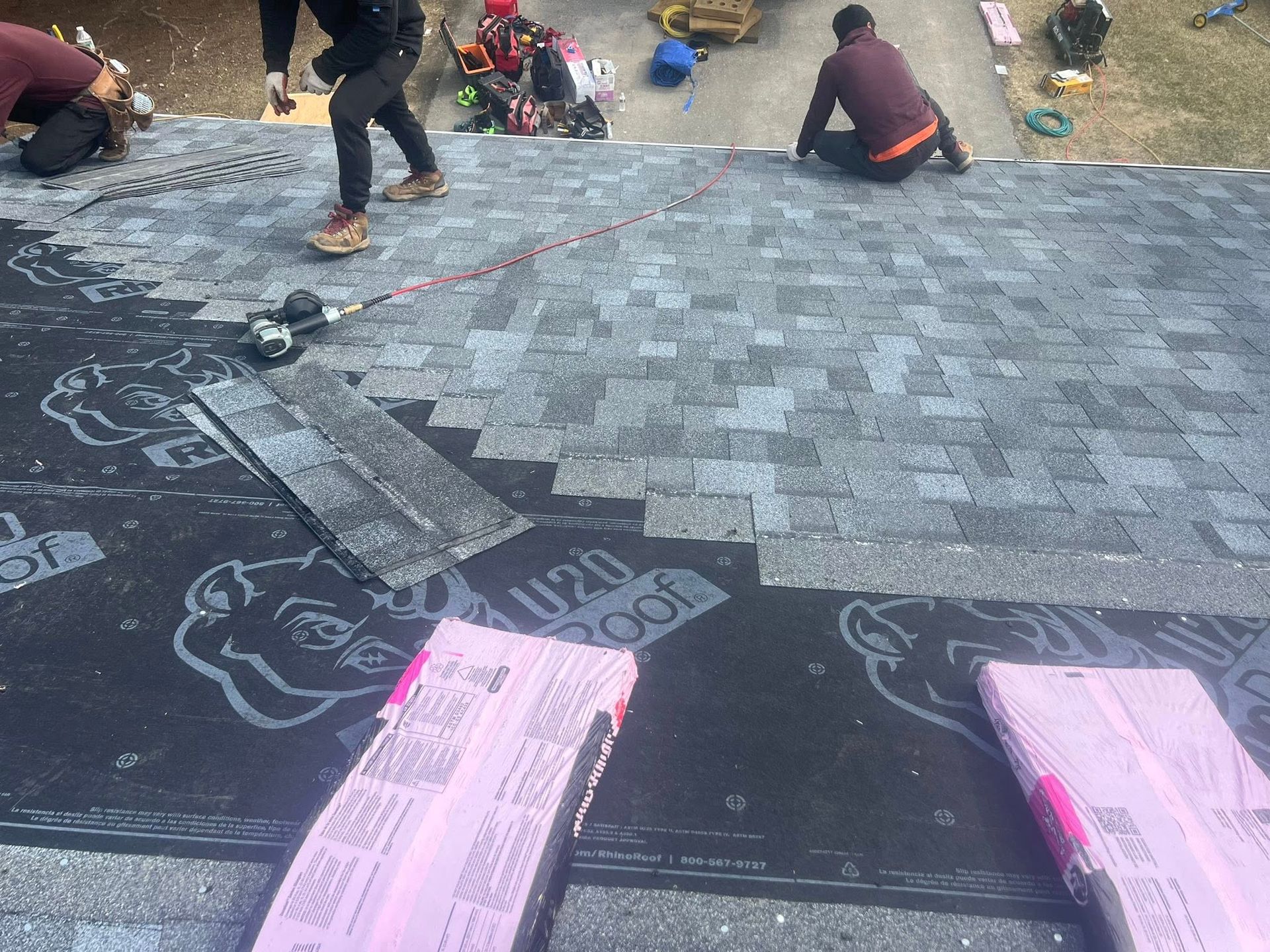 Workers laying black roofing shingles on a roof deck, with pink material in the foreground.