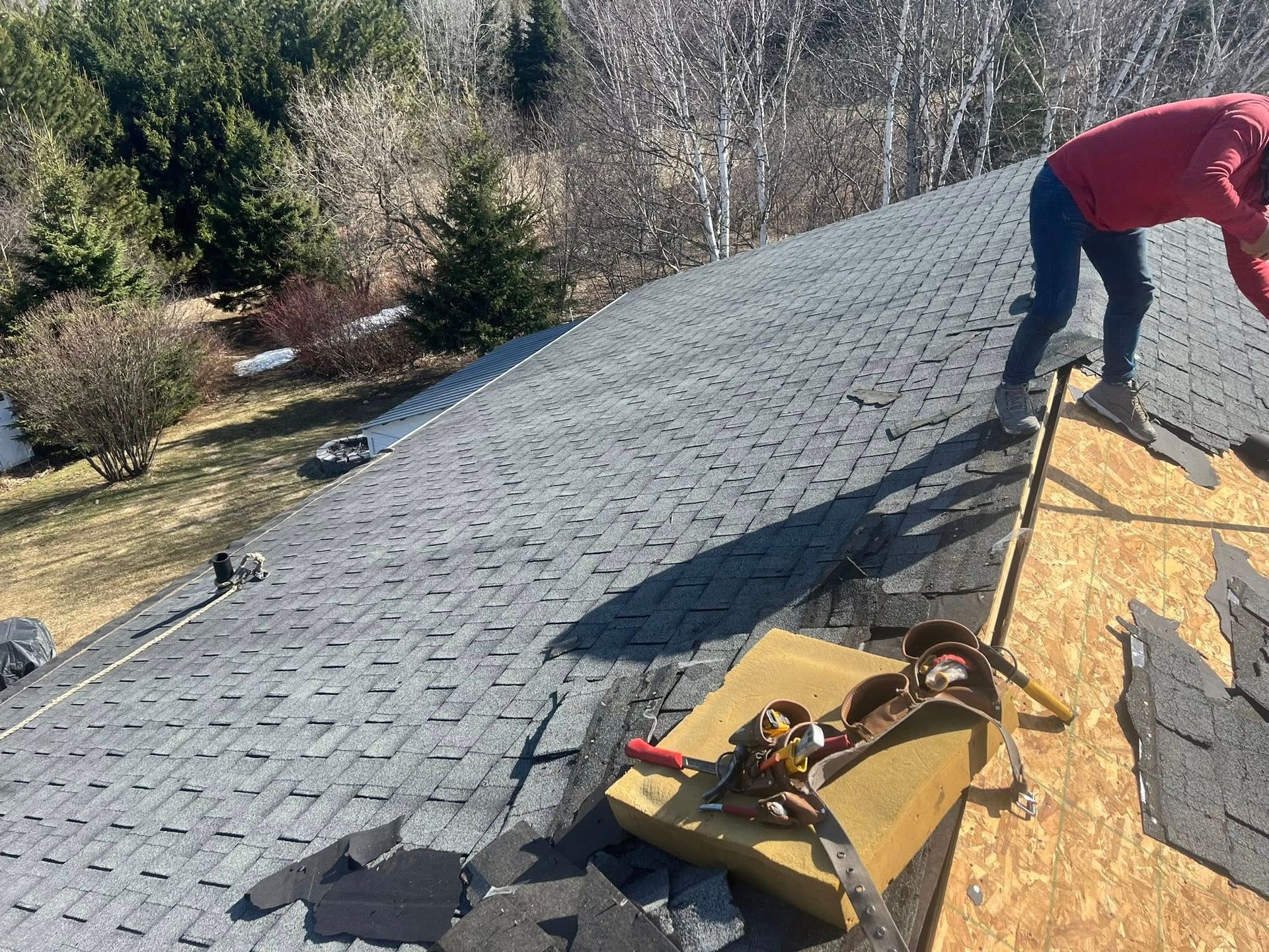 Worker installing gray shingles on a roof, with roofing materials scattered nearby.