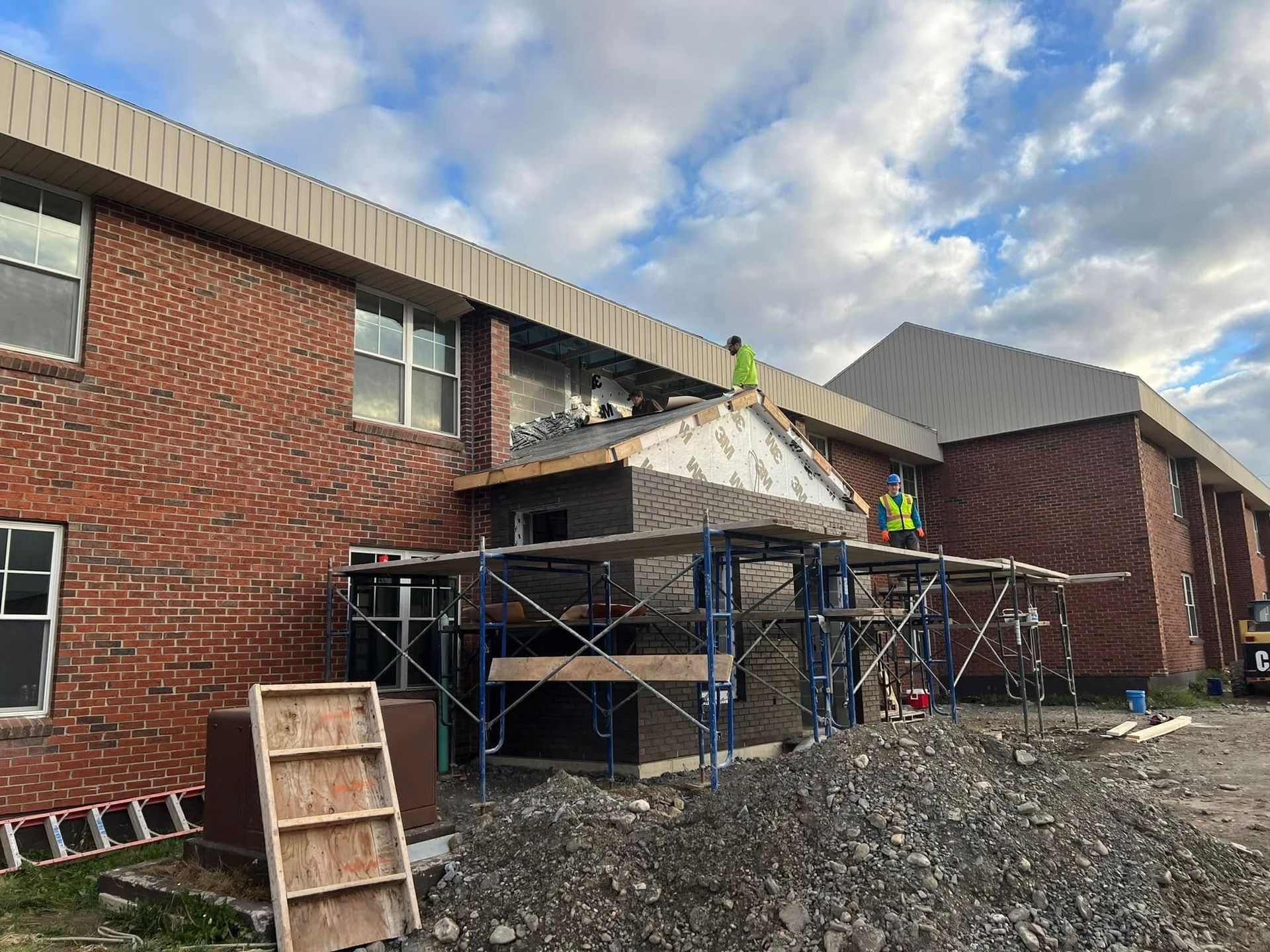 Scaffolded roof repair on a brick building with workers and piles of rubble under a cloudy sky