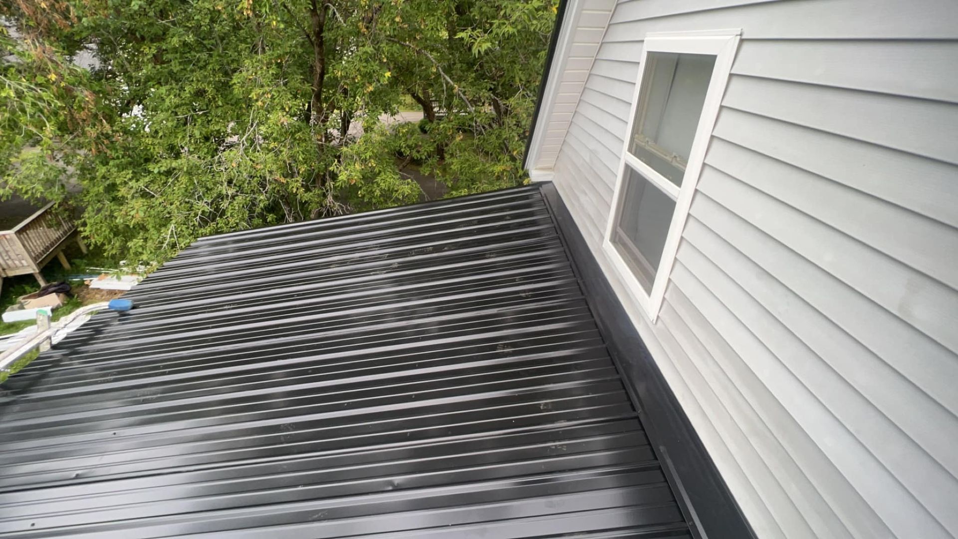 Black corrugated metal roof beside a gray house with a window, viewed from above