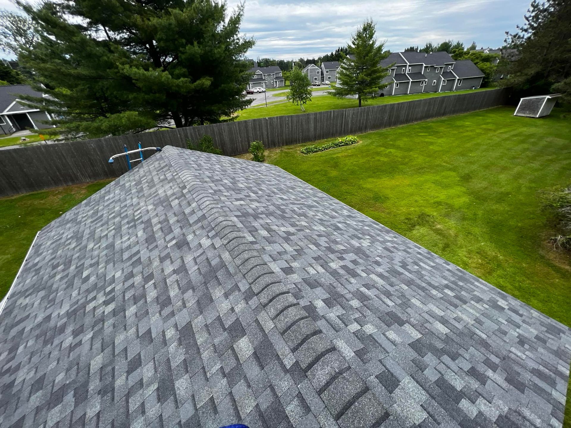 View from a shingled roof over a green backyard with trees and neighboring houses beyond a fence