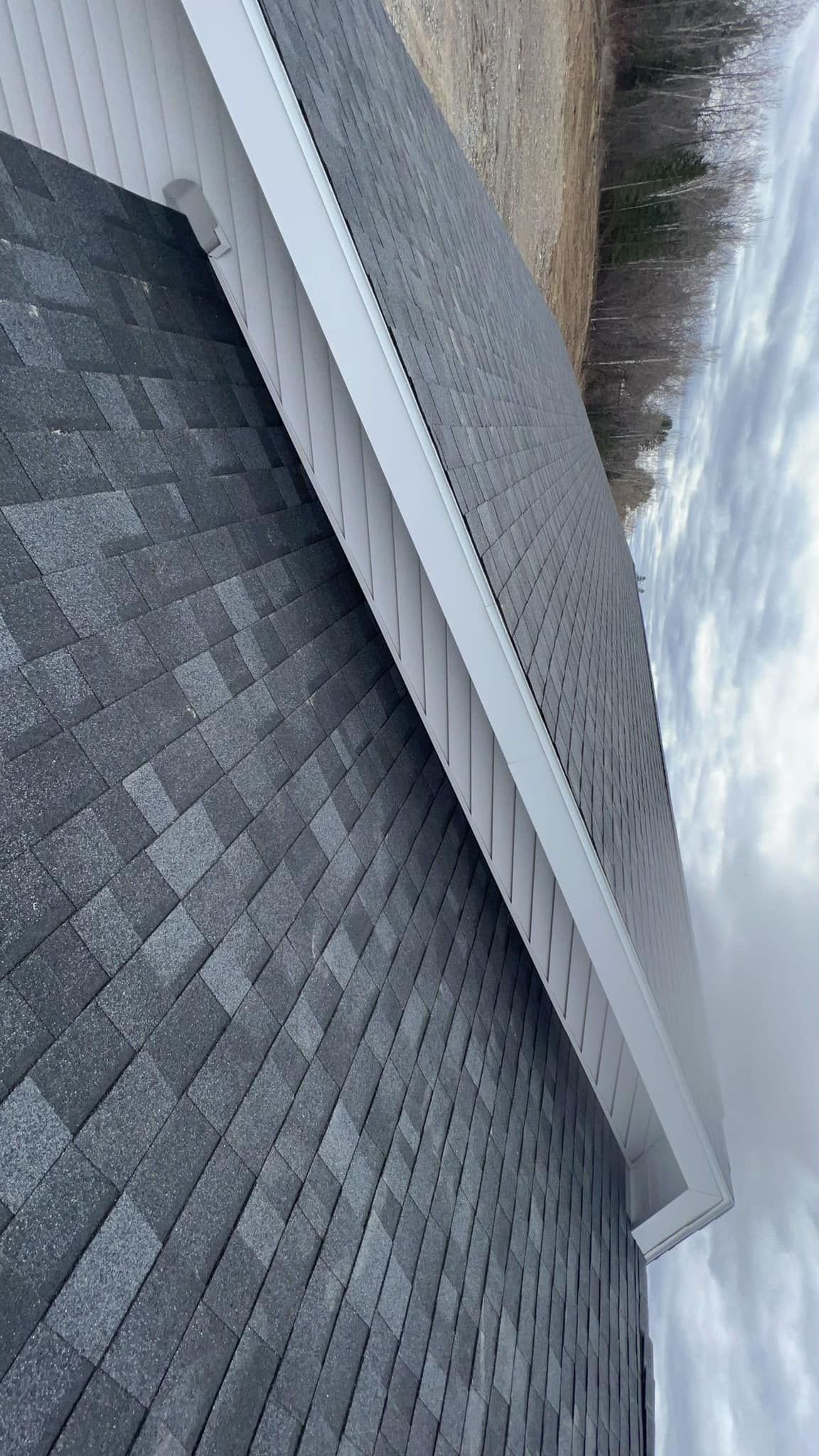 Close-up of dark gray shingled roof and white gutter beside a beige building under a cloudy sky