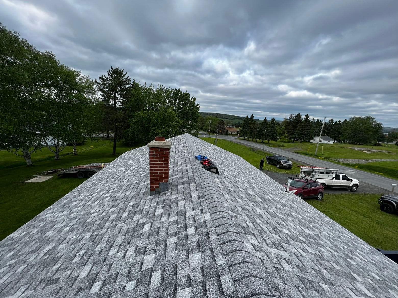 Rooftop view of a gray shingle roof with a brick chimney, cars below, and cloudy sky beyond