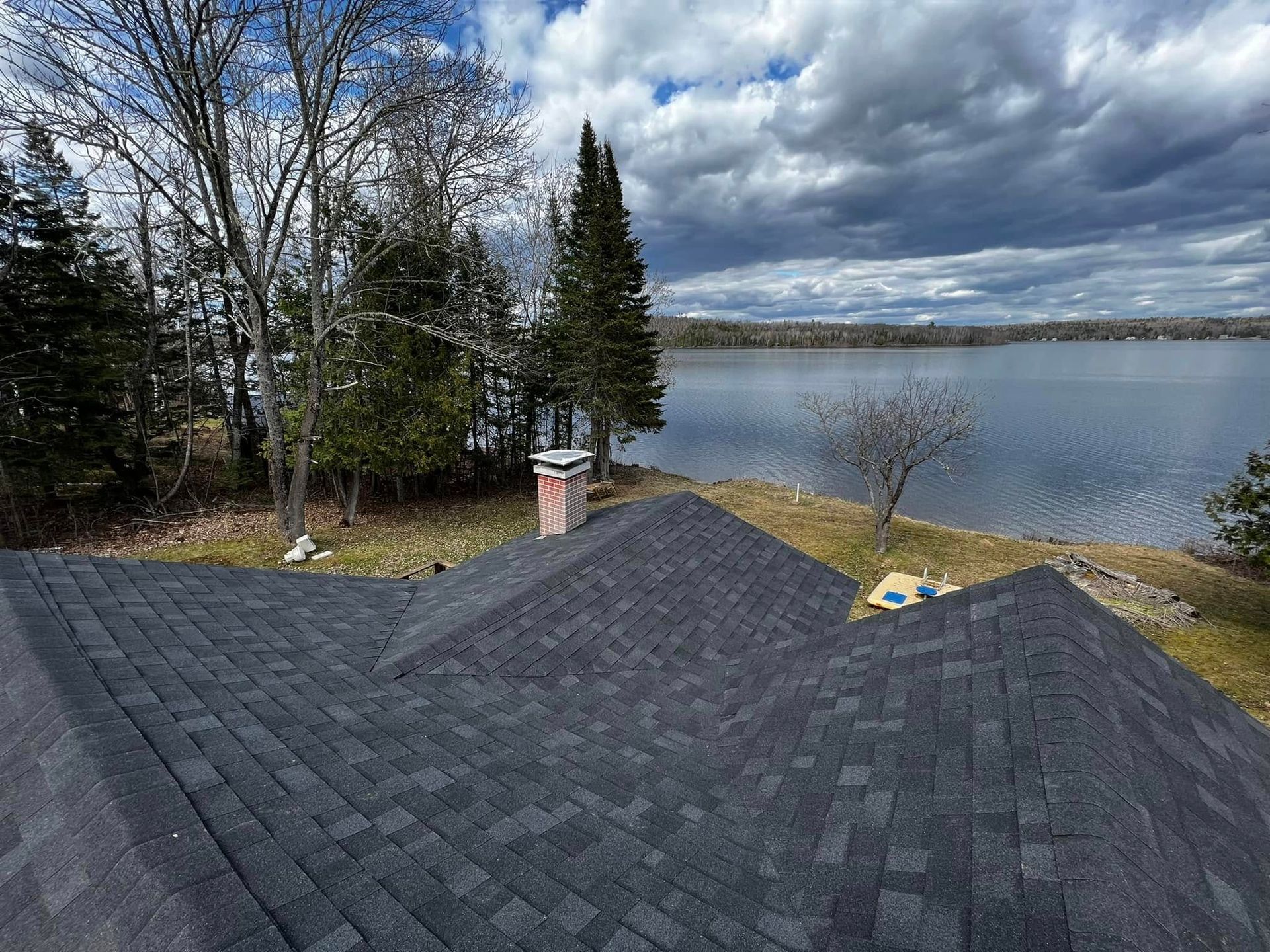 Dark gray shingled roof overlooking a lake under cloudy sky, with bare trees on the shore