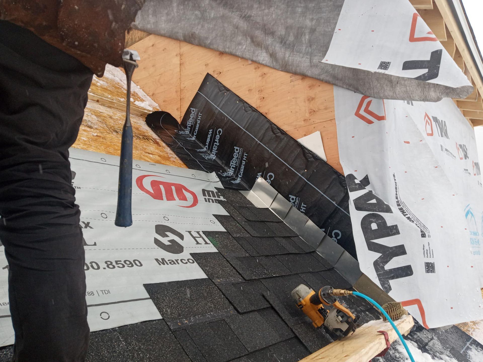 Roofing worker installing black shingles on a steep roof with tools and underlayment visible