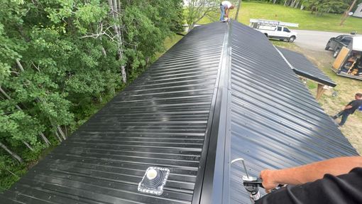 Roof viewed from ridge, with black metal panels and a person’s arm at the edge on a tree-lined property.
