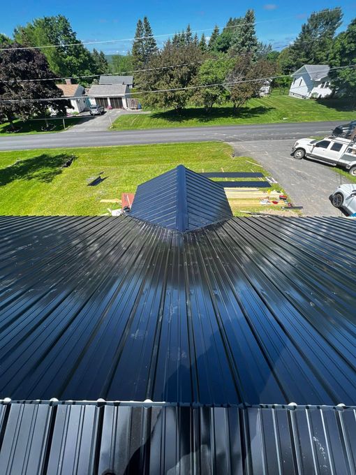 Rooftop view of a suburban street, with a dark metal roof, trees, houses, and a parked white truck.
