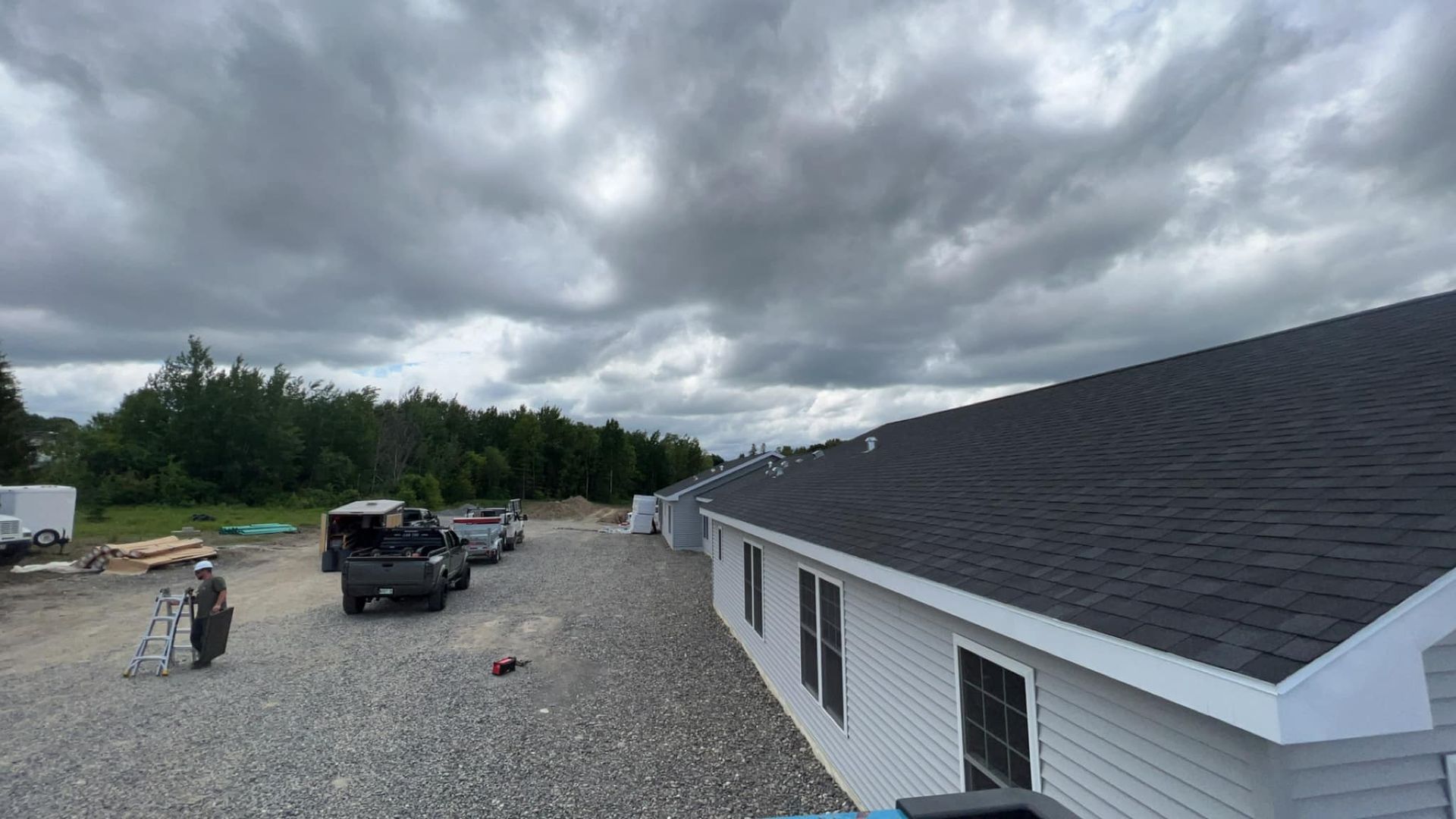 Gravel lot with parked vehicles beside a white building under dark cloudy sky