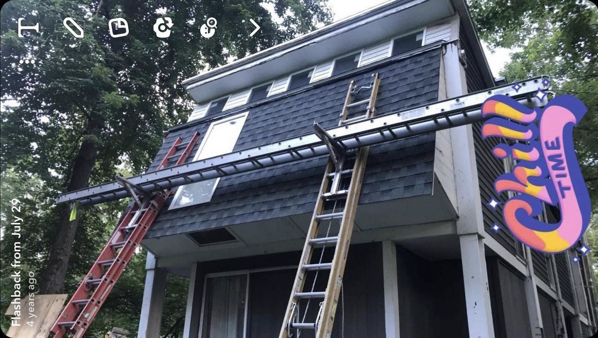Two ladders lean against a blue-gray house with a sloped roof, under construction in a wooded area.