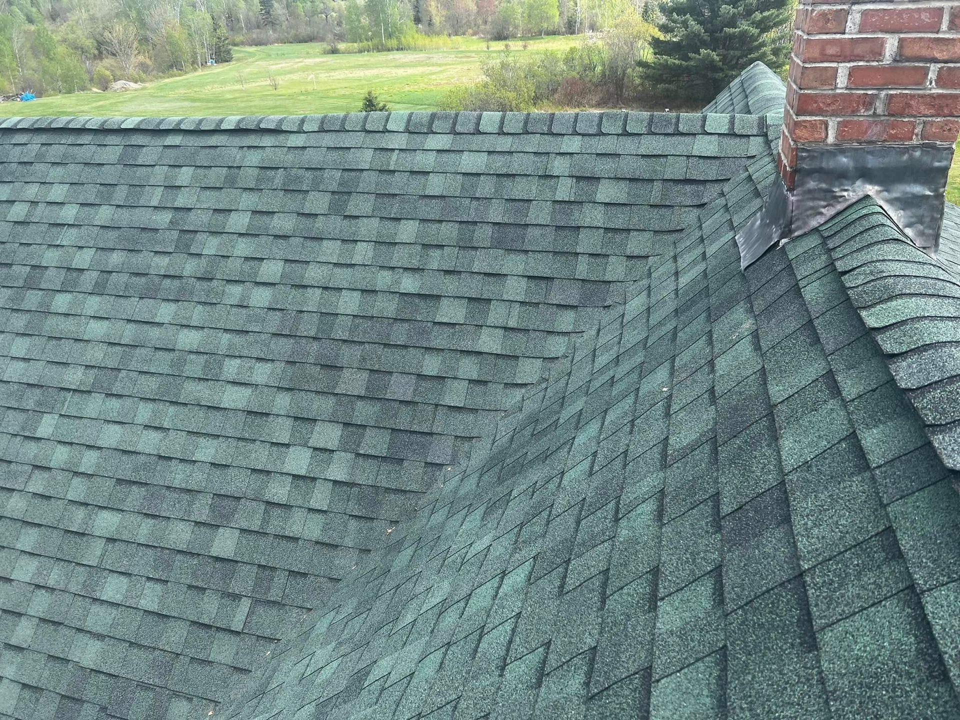 Green shingled roof with a brick chimney and a wooded landscape in the background