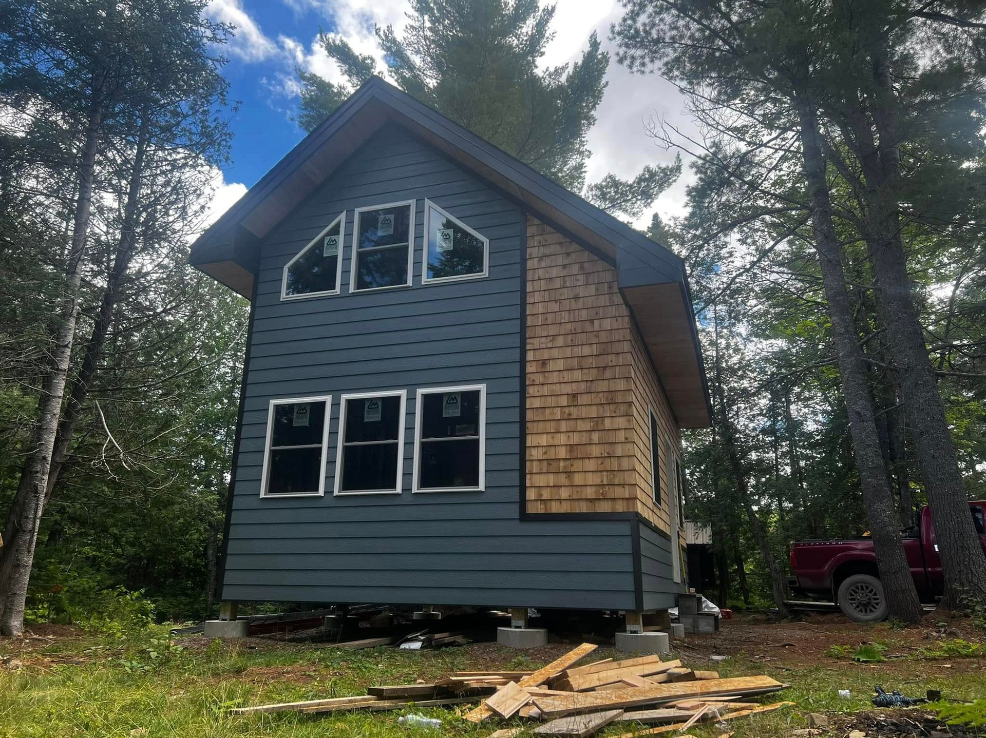 Small gray cabin under construction in a wooded clearing, with stacked lumber and a red truck nearby.