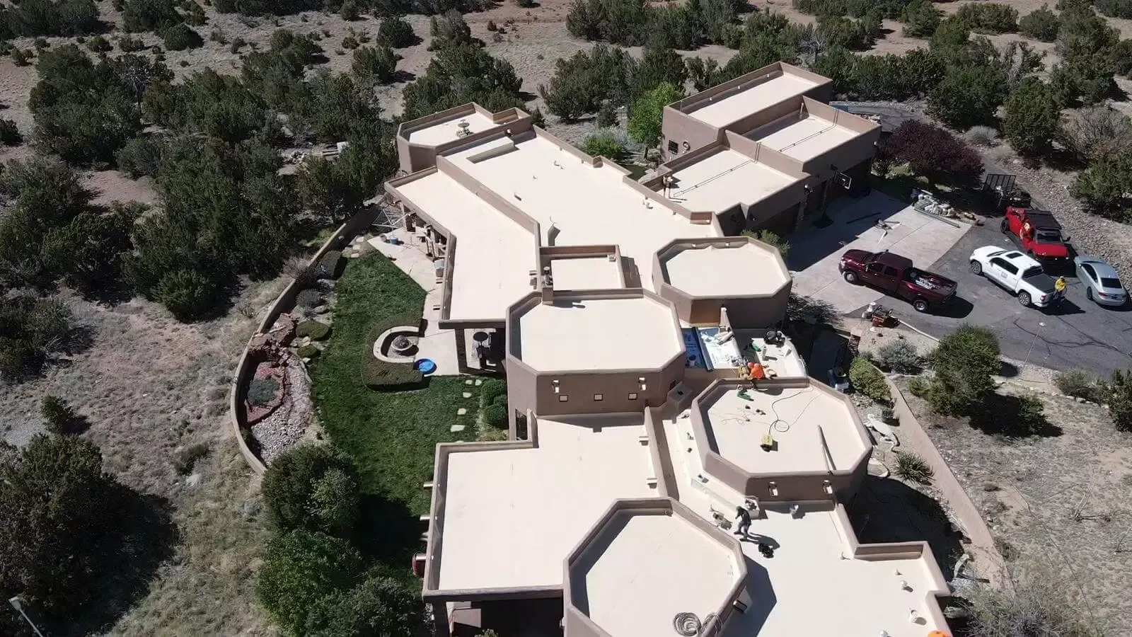 Aerial view of a large tan house in a desert landscape with parked cars and surrounding trees