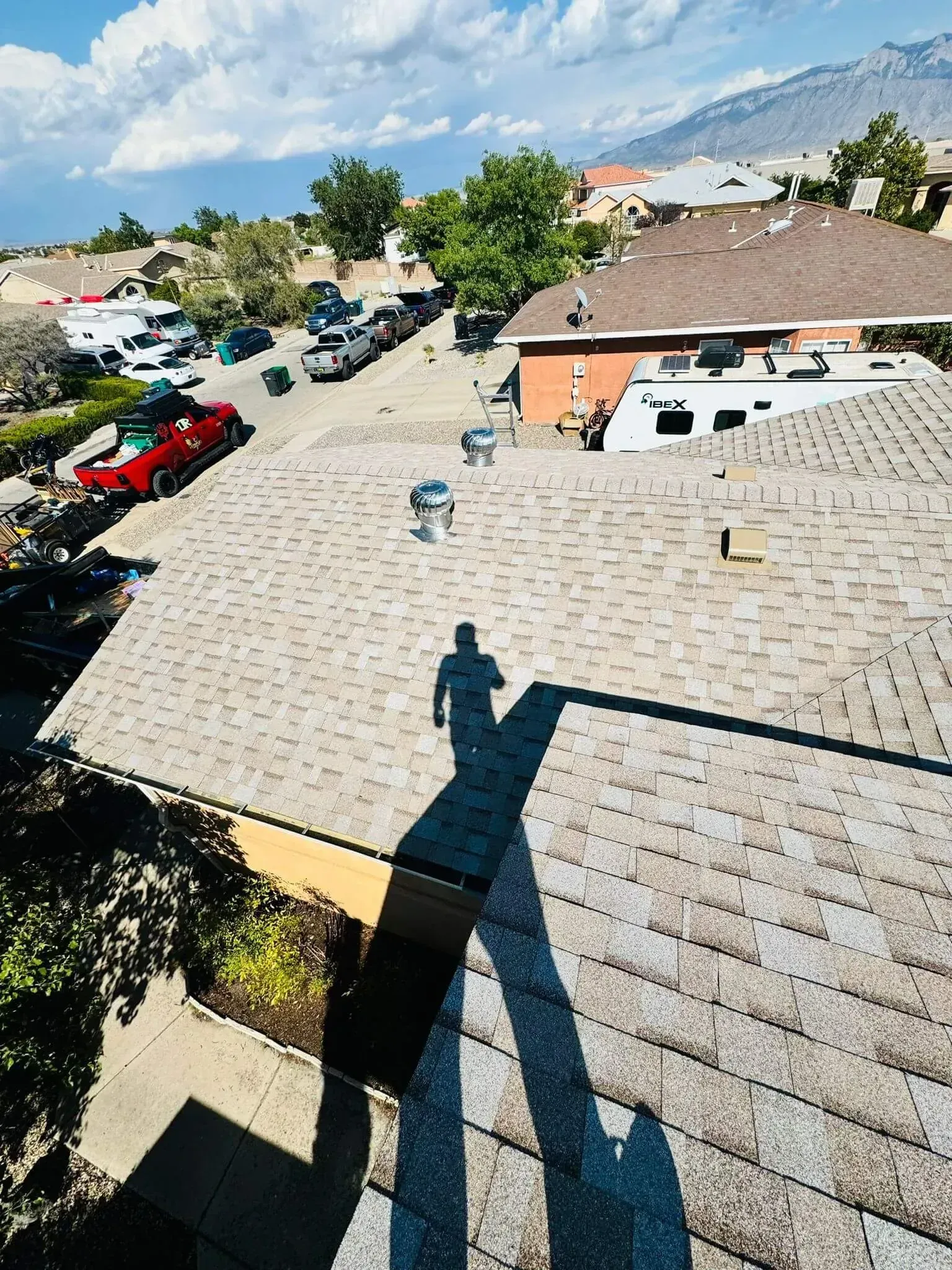 Rooftop with gray shingles and long shadow overlooking parked cars, trees, and neighboring houses under a blue sky