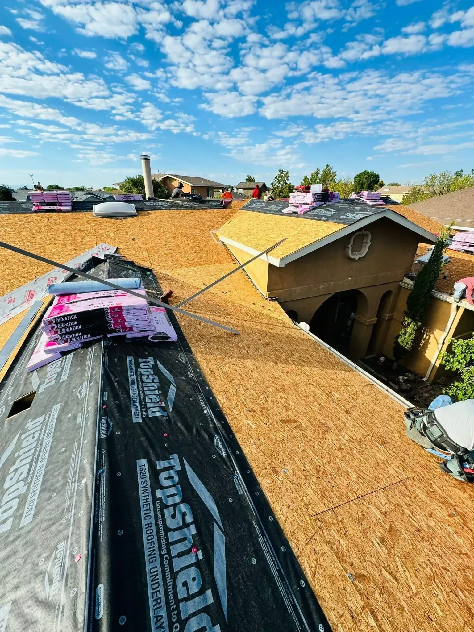 Roofing workers installing black underlayment on a tan shingle roof under a bright blue sky