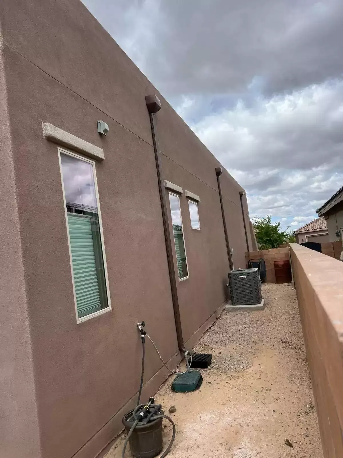 Narrow gravel side yard beside a brown stucco house, with windows, utility units, and cloudy sky.