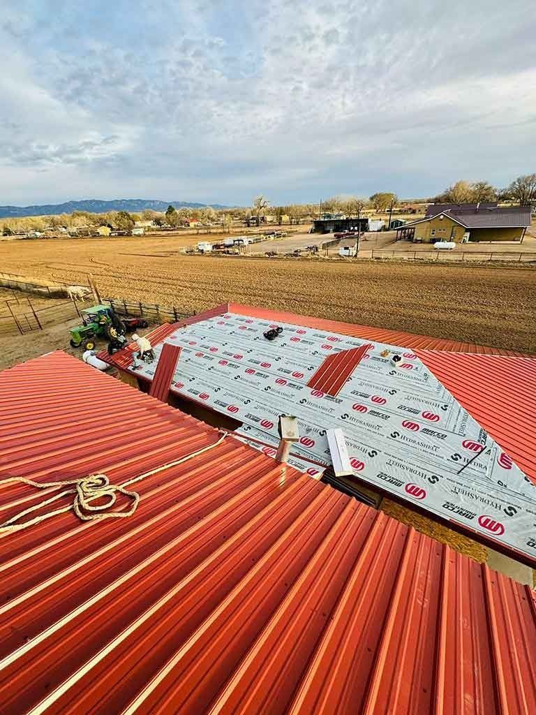Rooftop with red metal panels under construction, overlooking a rural field and houses beyond.