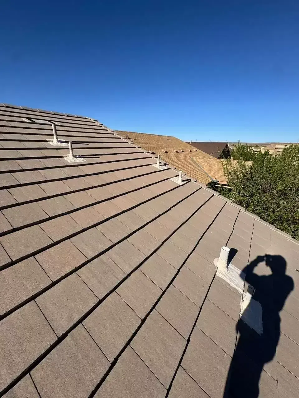 Roof shingles viewed from above, with a person’s shadow and blue sky over desert landscaping.