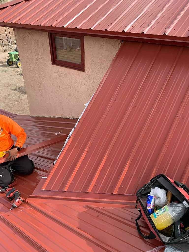 Worker installing red metal roofing on a house roof, with tools nearby.