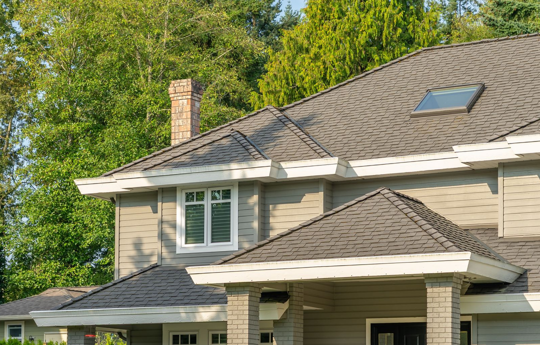 Two-story beige house with gray shingle roof and a dormer window, surrounded by green trees