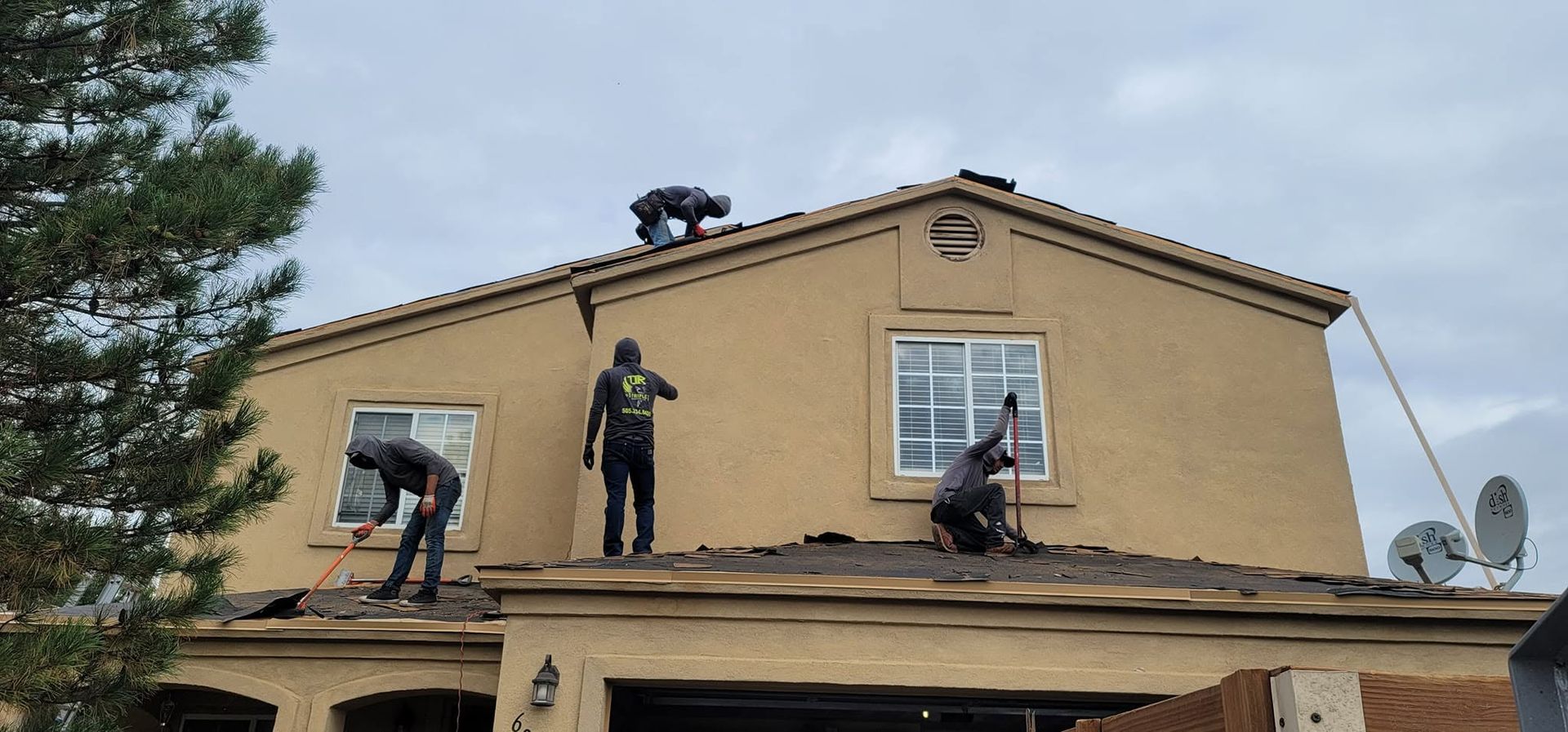 Three workers on a roof repairing a tan house, with ladders and a satellite dish visible.