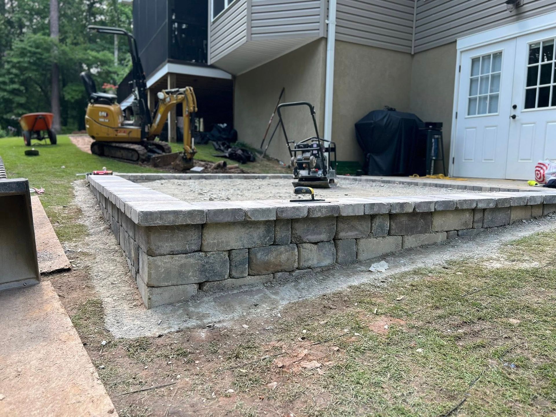 Stone retaining wall beside a house, with a small excavator and landscaping equipment in the yard