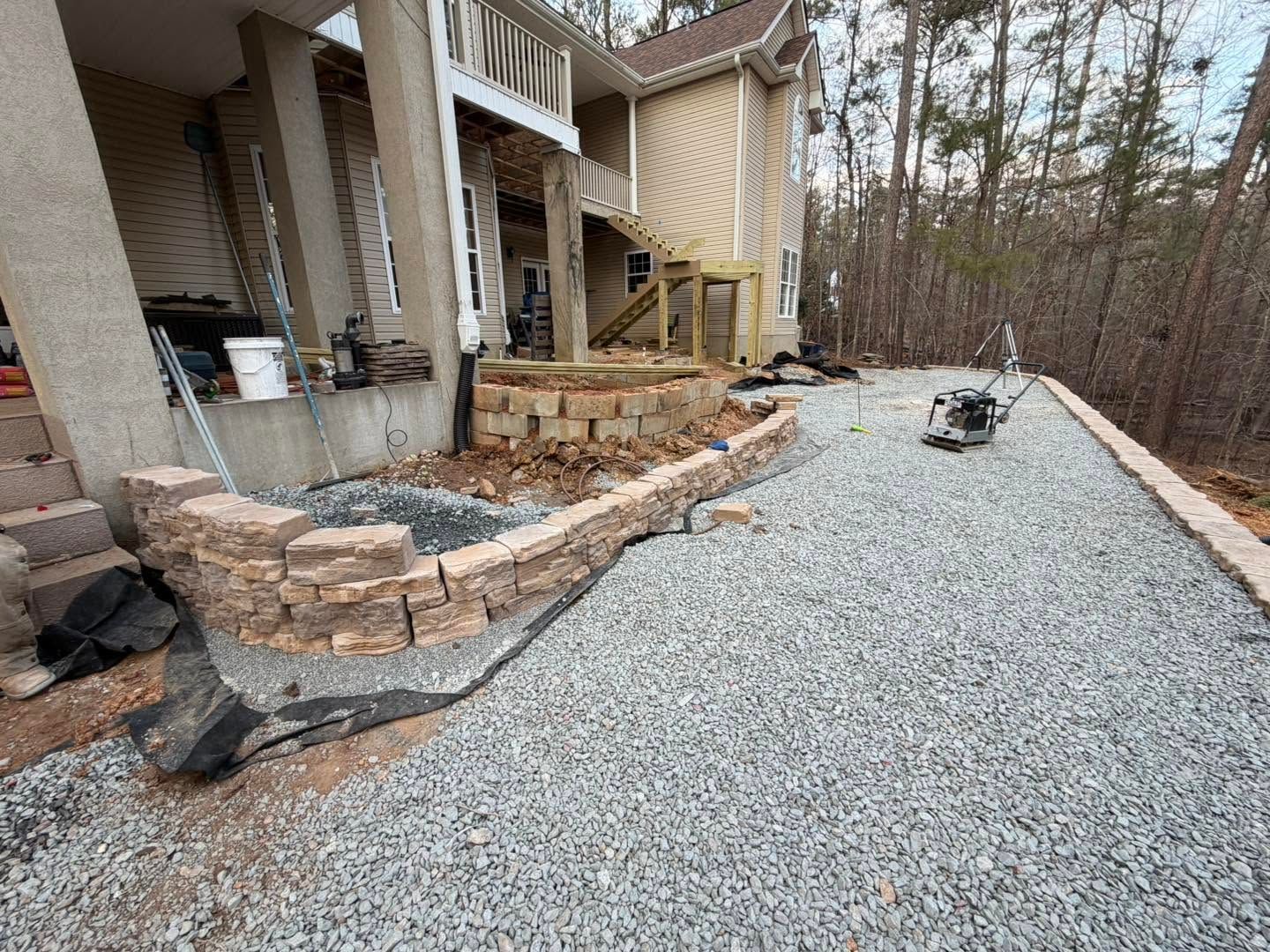 Stone retaining wall beside a gravel driveway under construction next to a house and wooded area