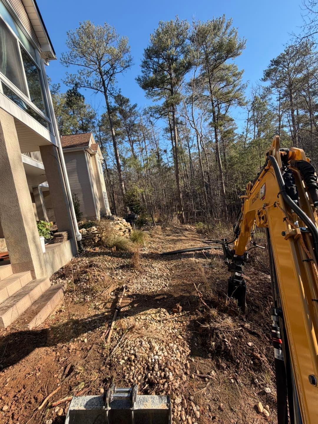 Excavator clearing brush beside a house in a wooded yard on a sunny day