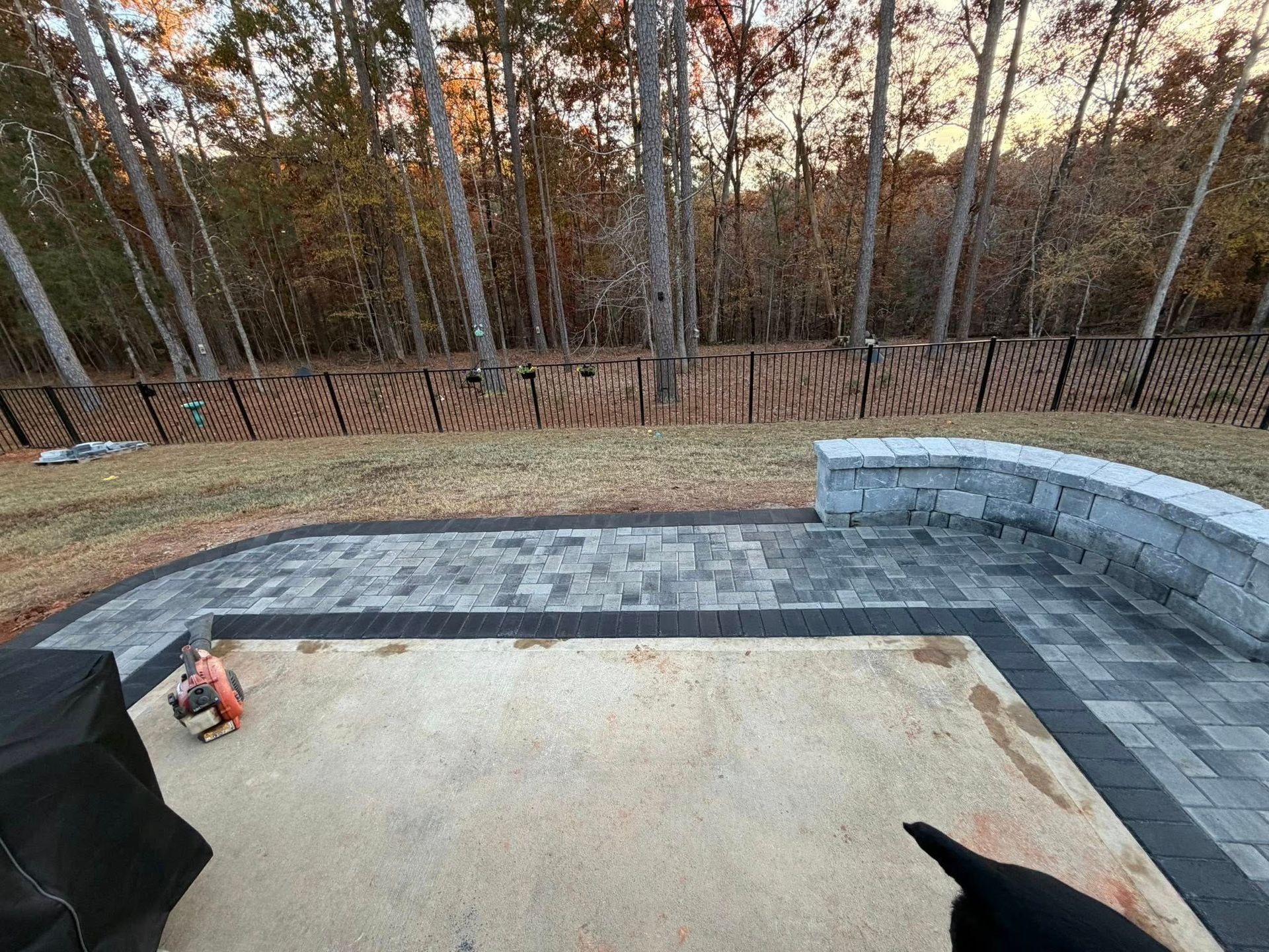 Backyard patio with gray stone seating wall, fire pit area, and wooded view beyond a black fence