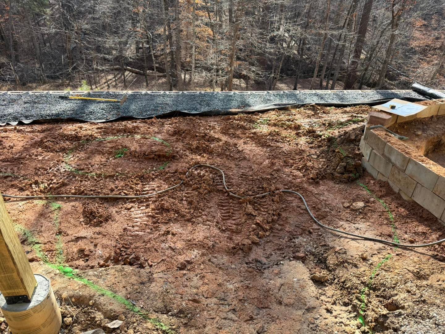 Bare dirt backyard with a black drainage tarp, cinder block wall, and wooded slope in the background