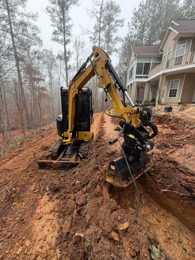 Excavator on muddy slope beside a house, clearing earth near a wooded lot.
