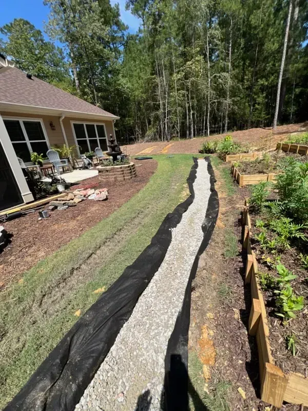 Narrow garden trench lined with black fabric and gravel beside a house and wooded yard