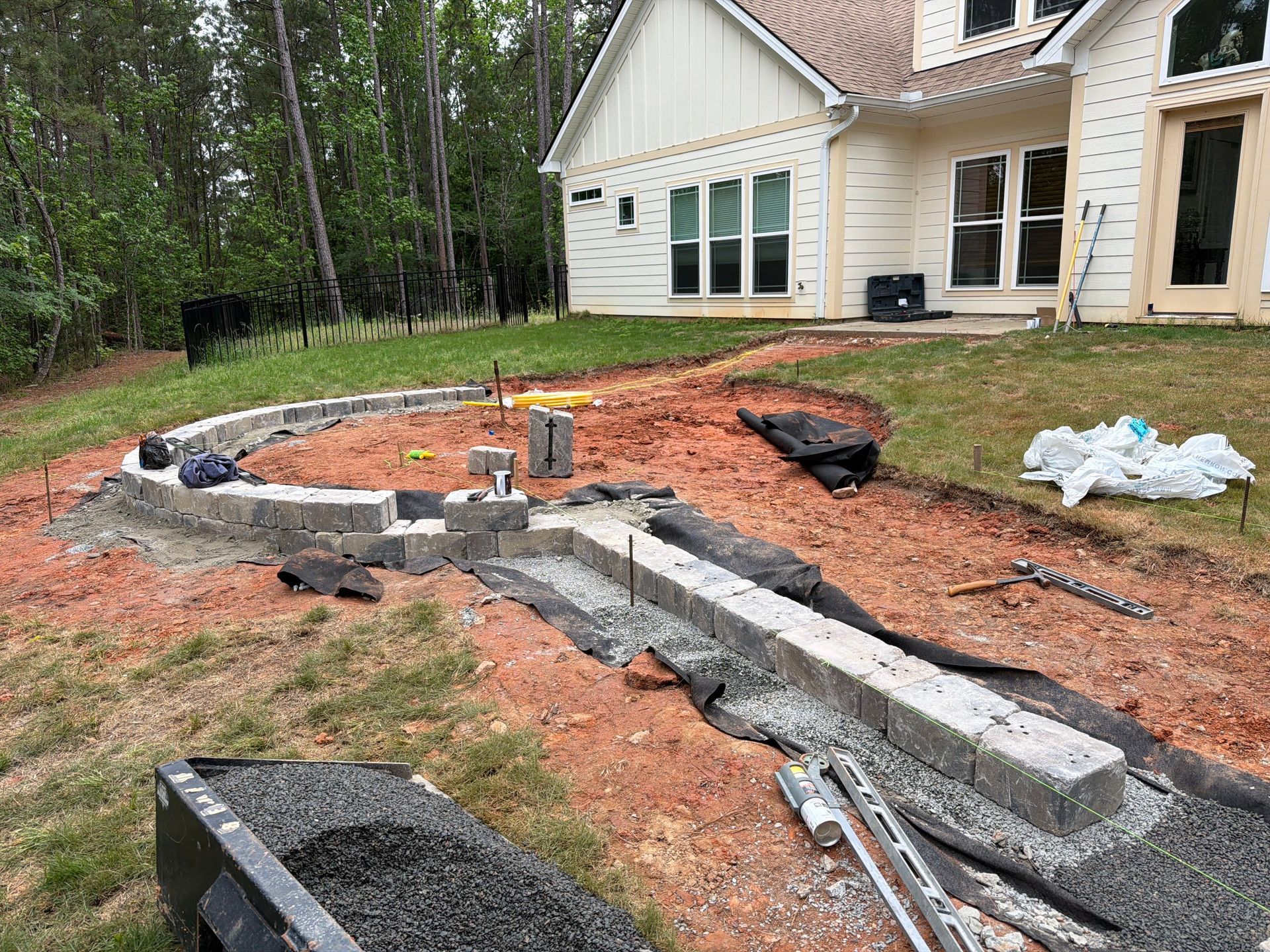 Curved cinder block foundation under construction beside a house, with dirt, tools, and materials scattered շուրջ the yard