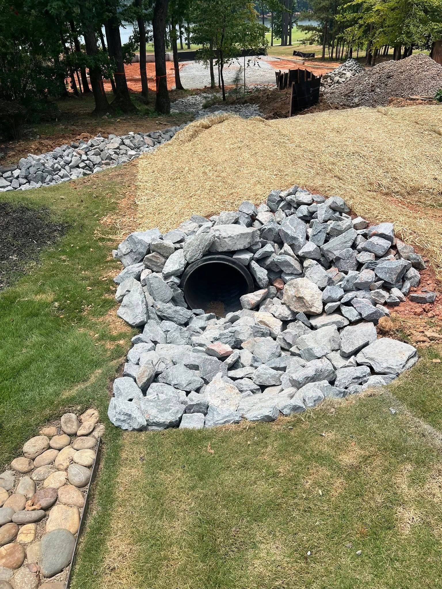 Stone-lined drainage channel in a grassy yard, with gravel and a culvert opening under trees.