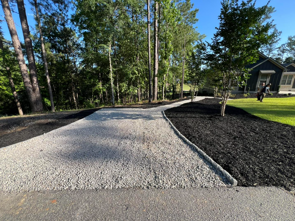 Gravel driveway beside a landscaped yard with trees and a house in the background