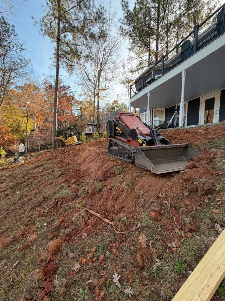 Excavator on a steep dirt slope beside a house, clearing soil after a landslide.