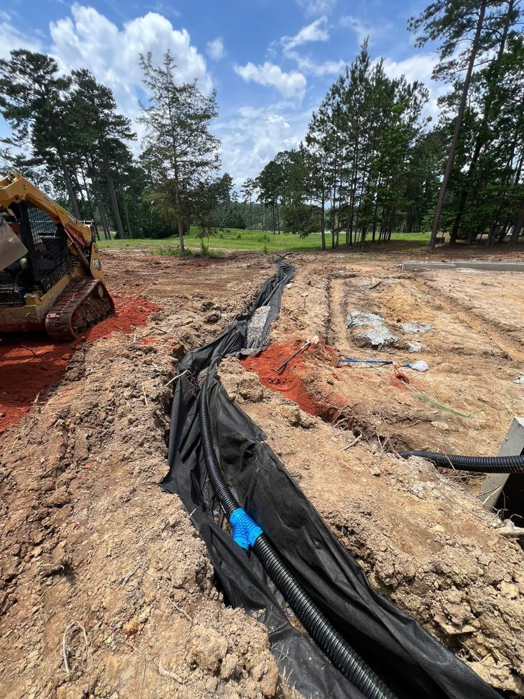 Excavator digging a trench with black utility conduit in a muddy construction site near trees