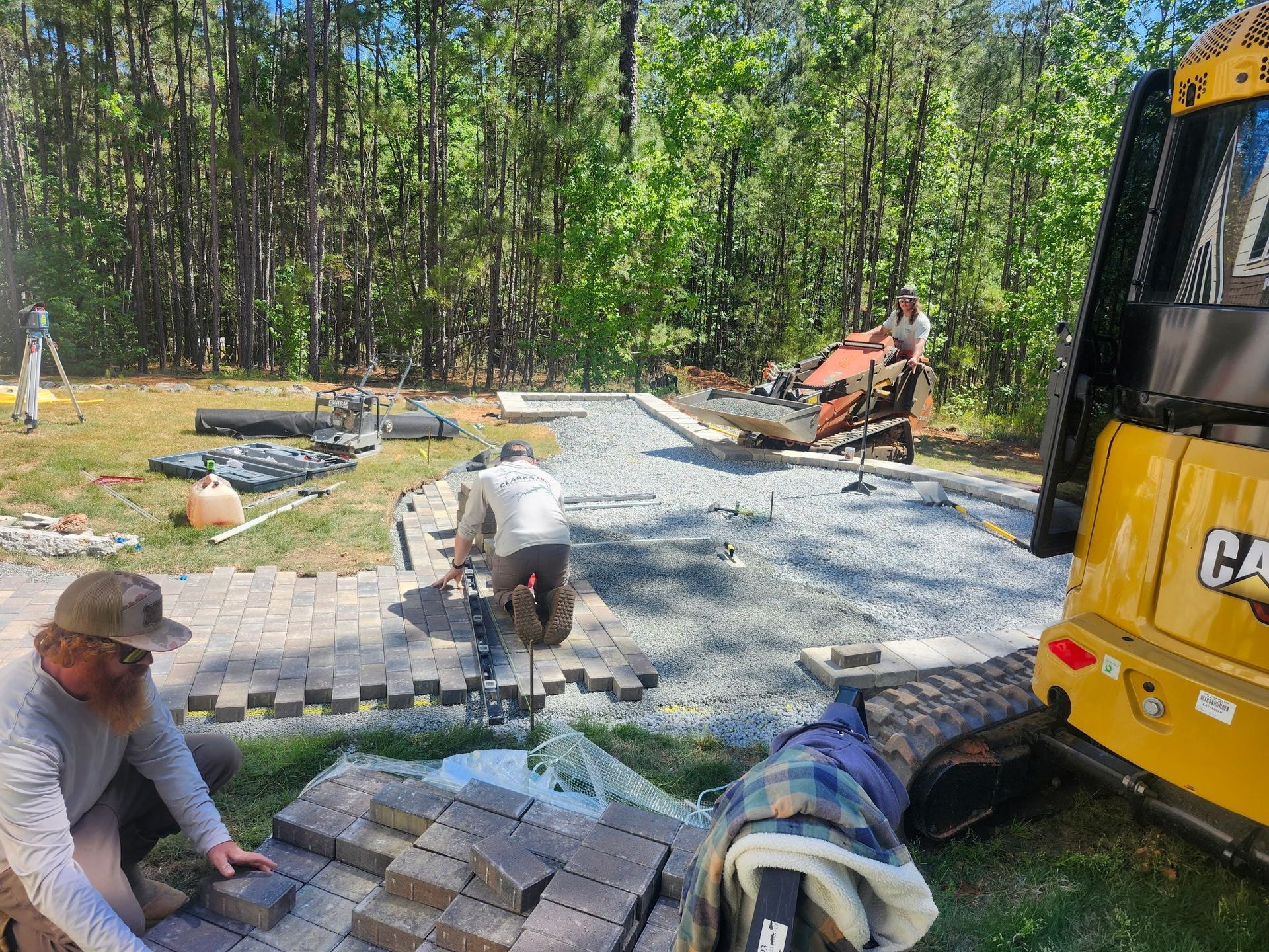 Workers laying pavers on a gravel patio beside a yellow excavator in a wooded yard