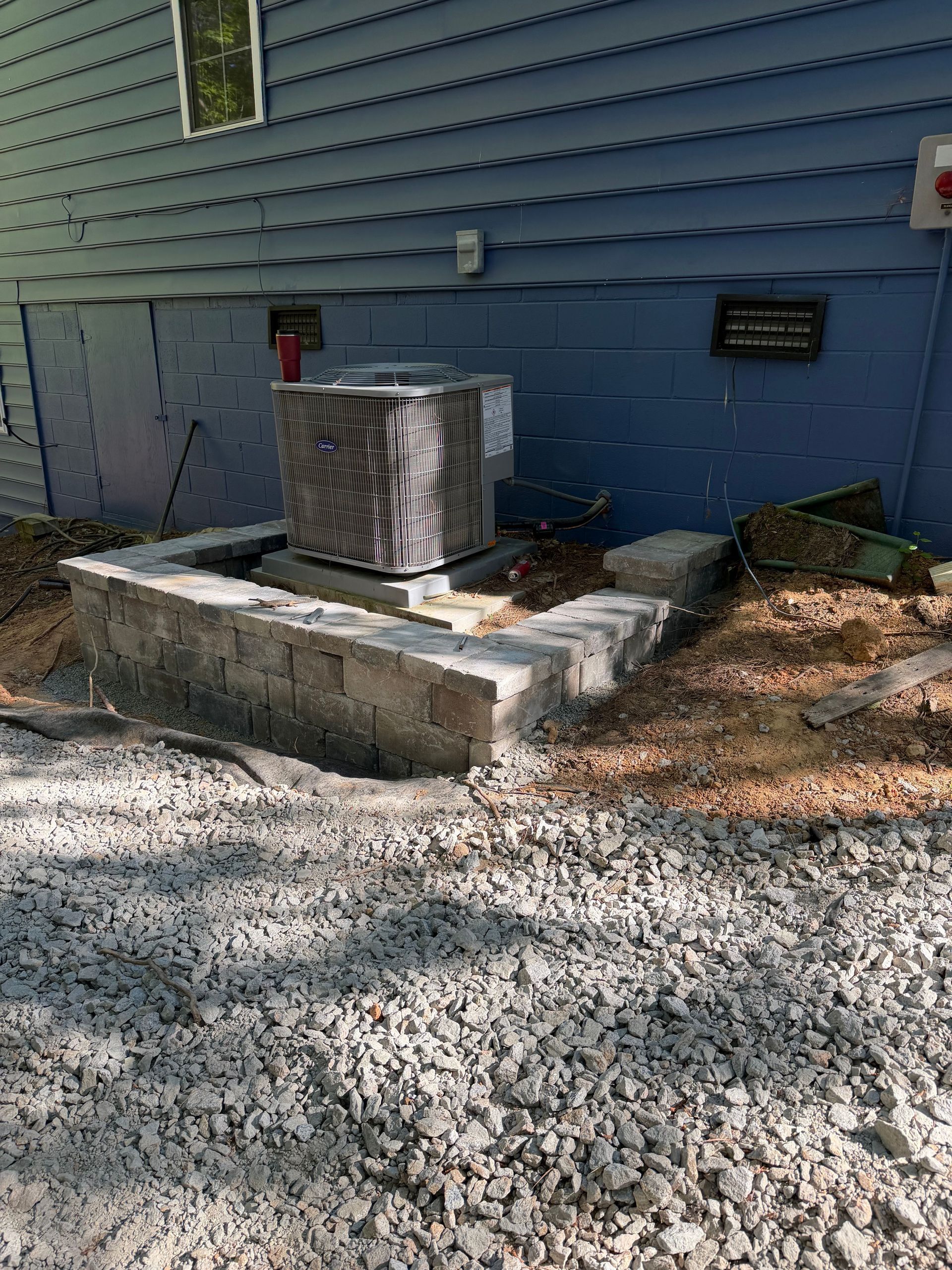 Outdoor HVAC unit on a concrete pad beside a blue house with gravel ground and fallen leaves.