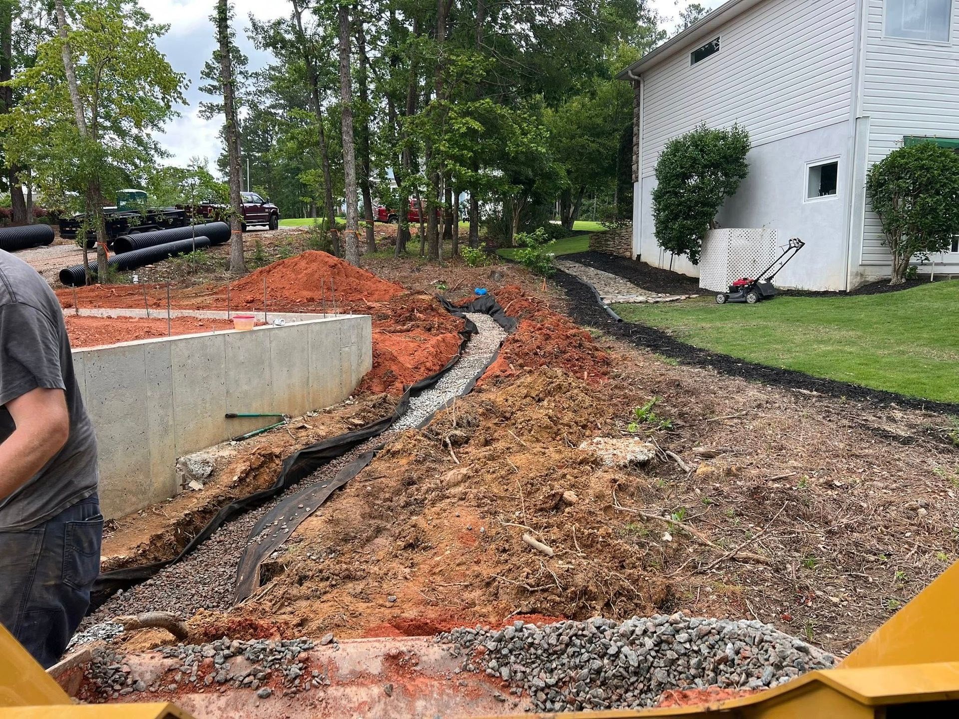 Backyard drainage trench under construction beside a white house, with gravel, dirt, and orange caution fencing