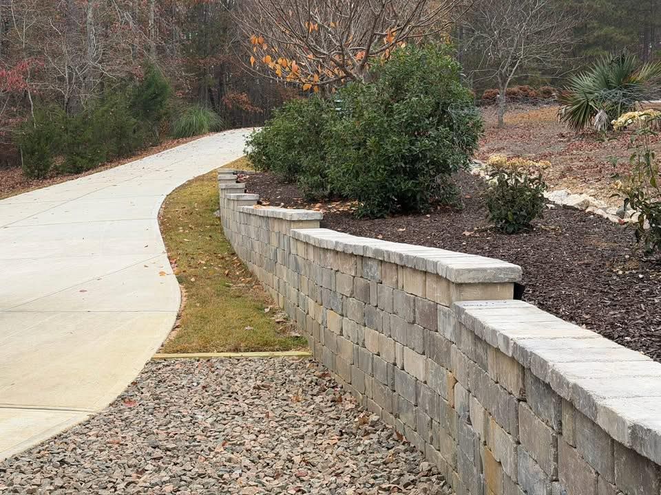 Curved concrete walkway beside a low stone retaining wall and landscaped shrubs in a yard