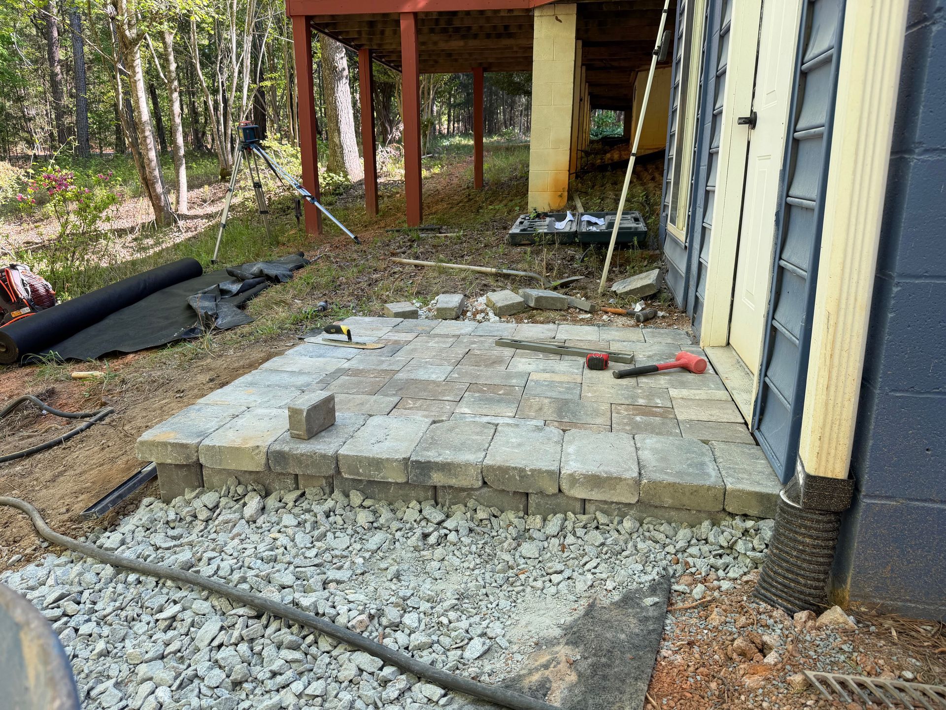 Partially built stone patio beside a house, with gravel, tools, and a wooded yard nearby.
